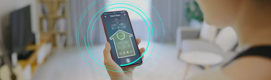 A hand holding a smartphone running a home-control app, with a living room behind it