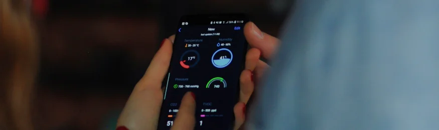 A hand holding a smartphone that shows an IoT dashboard with device readings