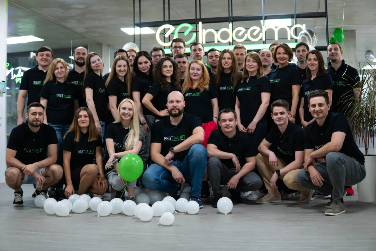 Group photo of the Indeema team in black BEST MOOD shirts in front of a lit Indeema sign, with balloons on the floor