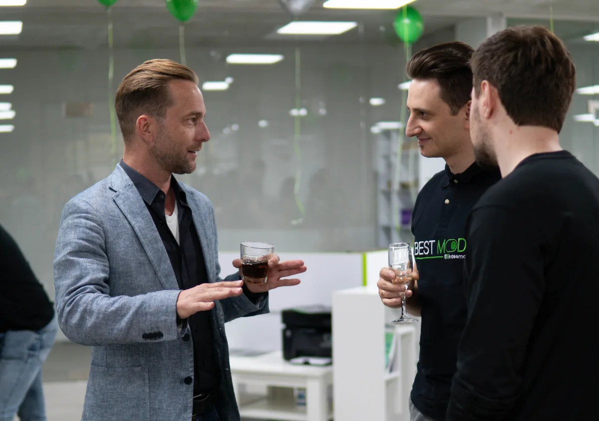 Man in a grey blazer talking with two men, one in a BEST MOOD shirt, at an office party with green balloons