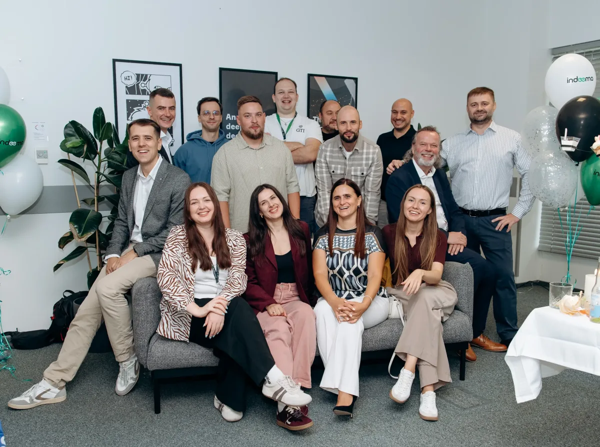 Group photo of about fifteen people on and around a sofa, with Indeema-branded balloons in an office