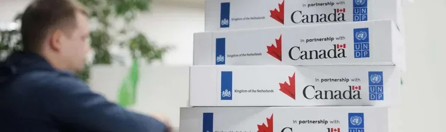 Stack of device boxes labelled "In partnership with Canada", Kingdom of the Netherlands and UNDP, a man at a desk behind