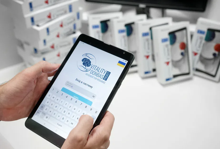 Hands holding a tablet showing the Vitality Donbas Project login screen, with boxed tablets in the background