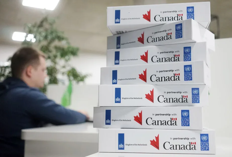 Stack of boxed devices labelled "In partnership with Canada", Kingdom of the Netherlands and UNDP, a man working behind