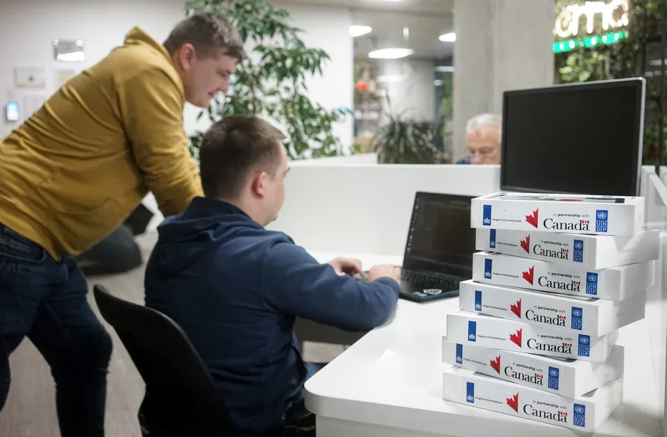 Two men at a laptop beside a stack of device boxes labelled "In partnership with Canada" and UNDP