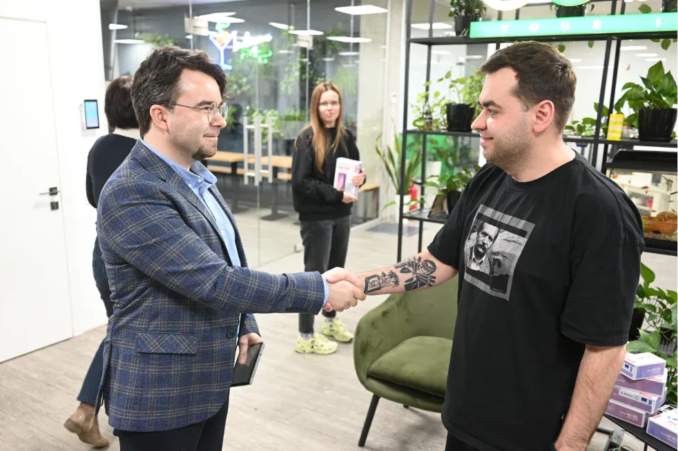 Two men shaking hands in an office while a colleague holds a boxed tablet in the background