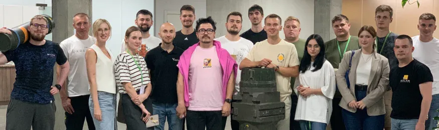 Group photo of about twenty people in an office, two of them holding olive-green equipment cases