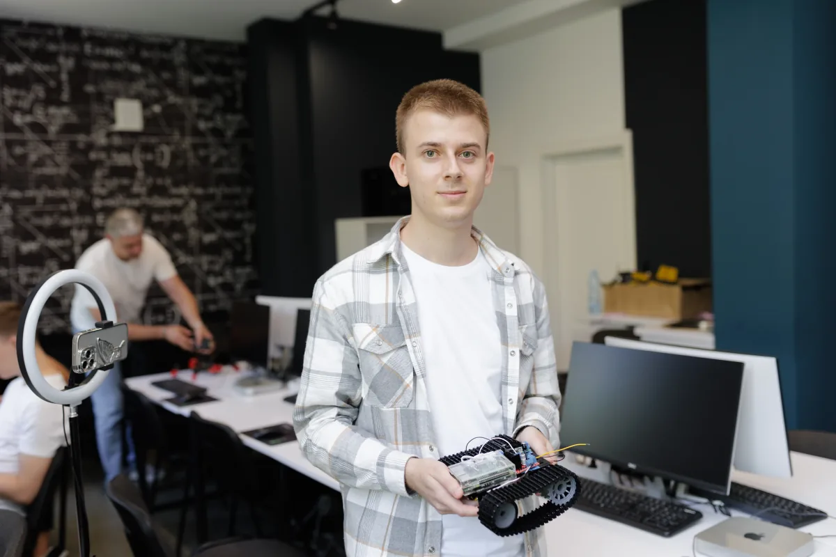 Student in a checked shirt holding a tracked robot chassis in the IoT lab, a man handling a drone in the background