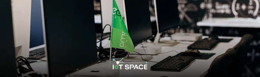 Darkened computer lab with monitors, keyboards and a green Indeema desk flag, with the IoT Space logo overlaid