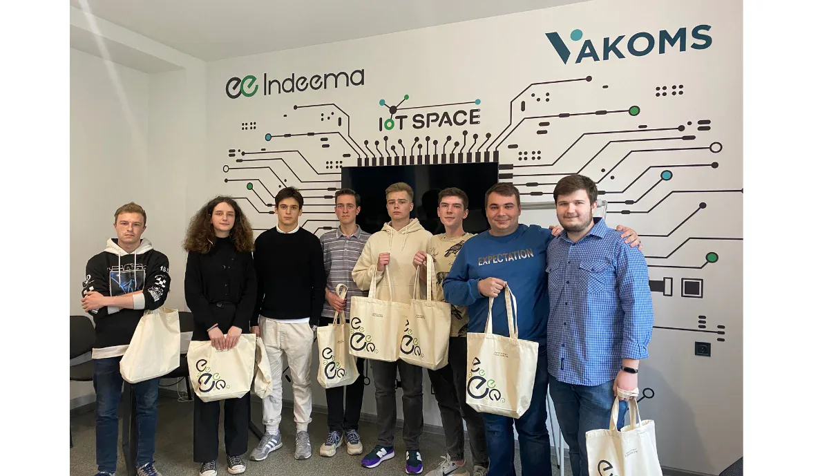 Group photo of IoT Challenge students and two hosts holding Indeema tote bags in front of the Indeema, Vakoms and IoT Space wall