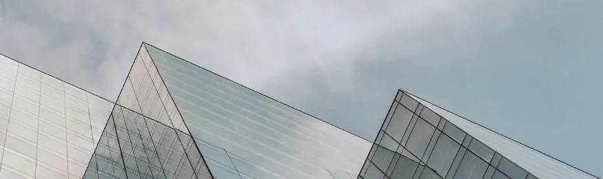 Upward view of angular glass office building facades against a cloudy grey sky