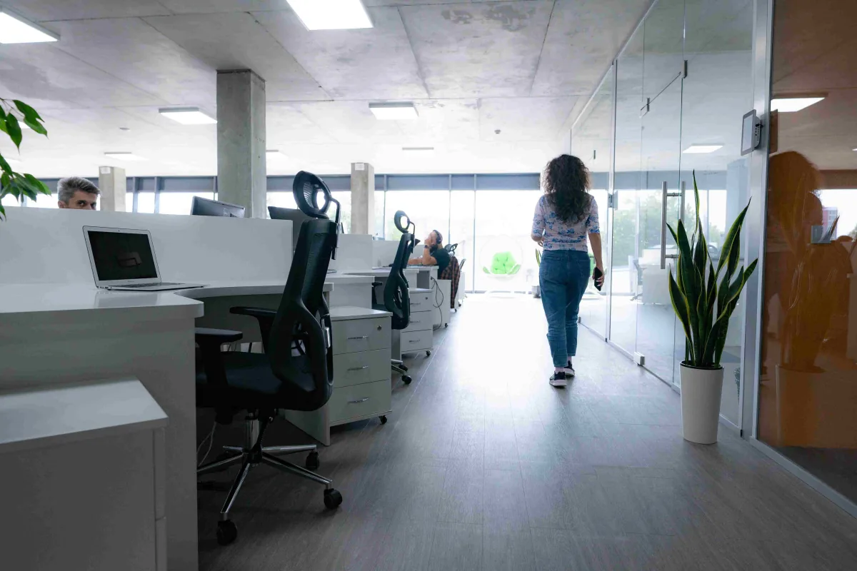 A woman walks down a bright office aisle between desks and glass-walled rooms toward large windows