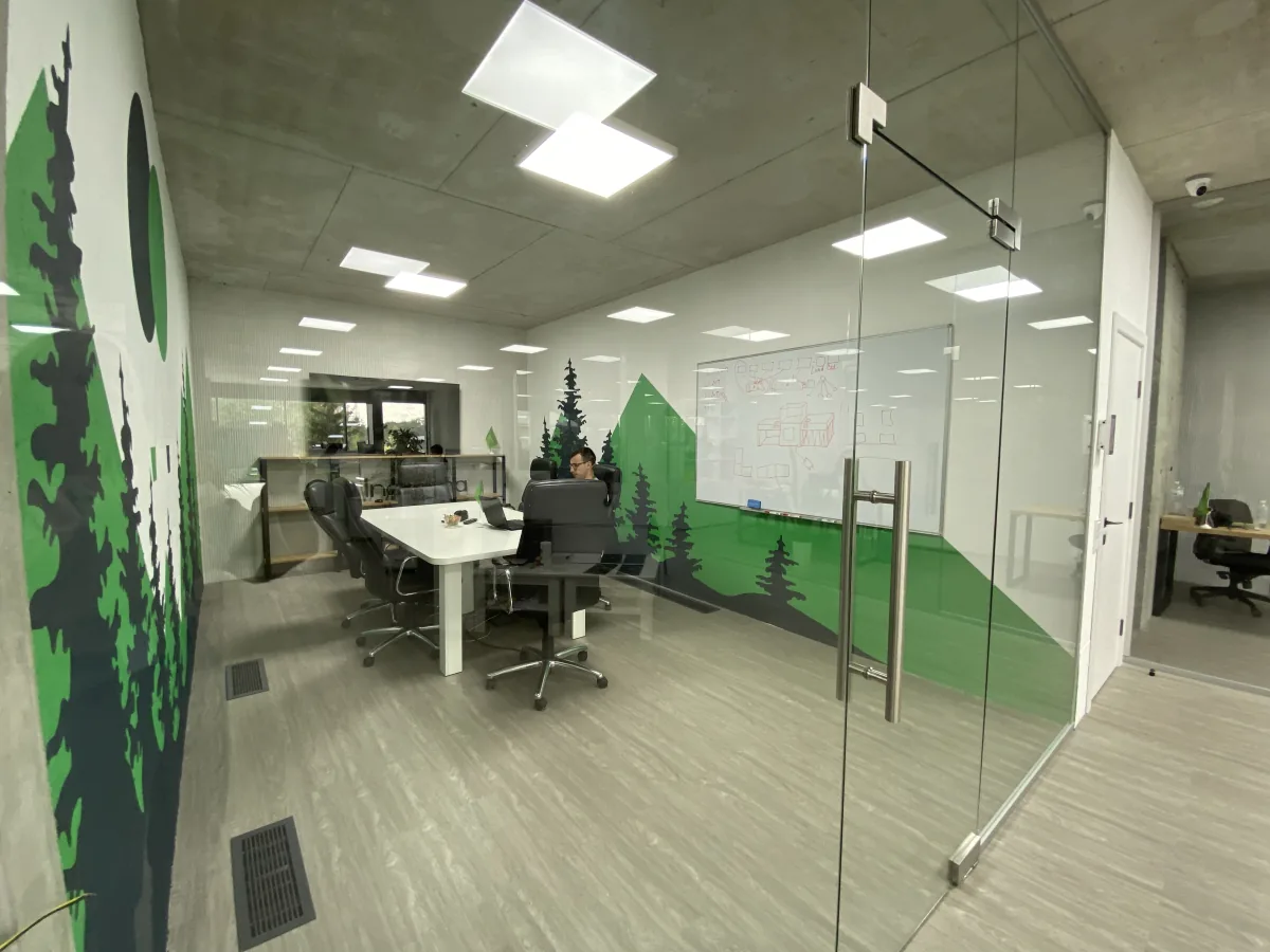 Glass meeting room with a forest mural, whiteboard and white conference table where a person works on a laptop