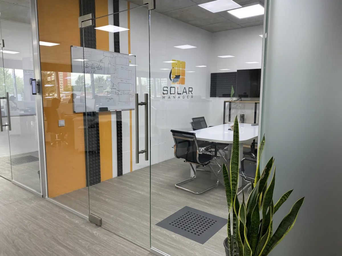 Glass-walled Solar Manager meeting room with a whiteboard, white conference table and a snake plant at the entrance