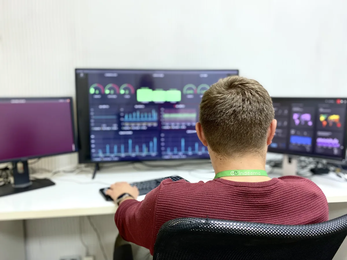 Engineer wearing an Indeema lanyard works at three monitors showing dashboards with gauges, bar charts and maps