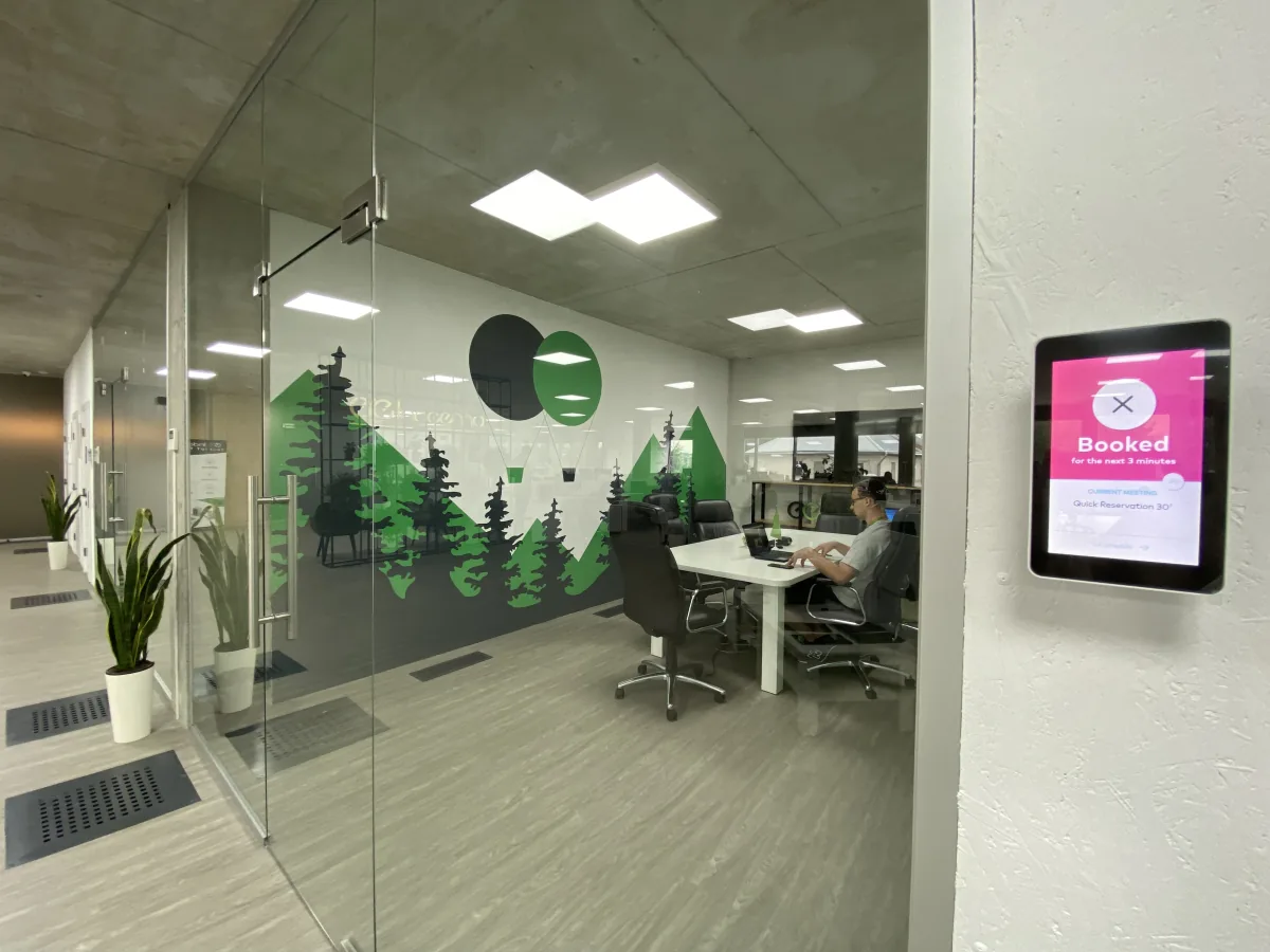 Meeting room with forest mural behind glass and a wall-mounted booking tablet showing Booked for the next 3 minutes