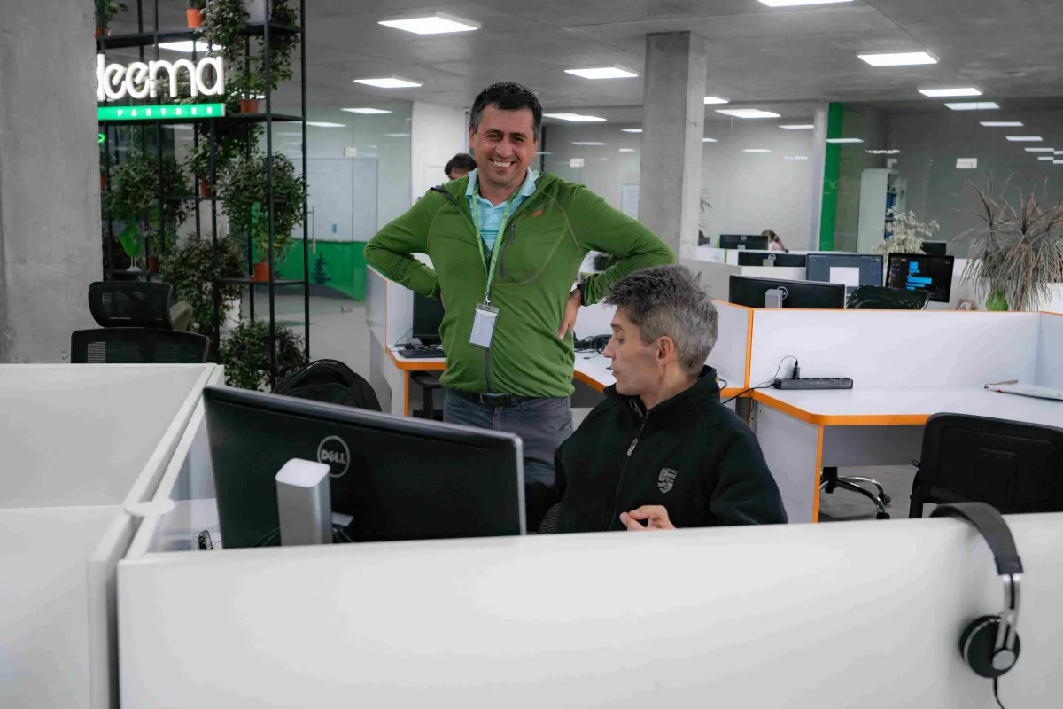 Two Indeema employees talk at a desk in the open-plan office, one standing and smiling, the other seated at a monitor