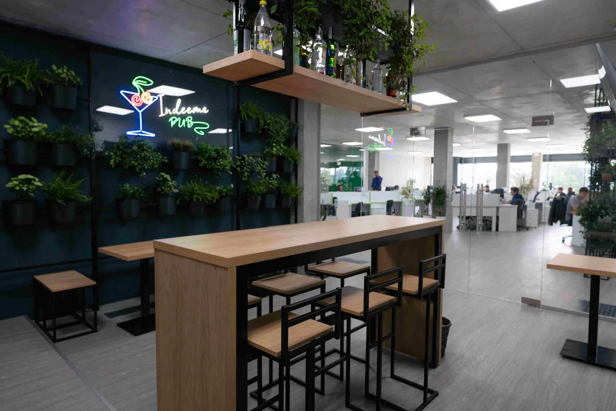 Office lounge with a neon Indeema Pub sign, plant wall, wooden bar table with stools and a view into the workspace