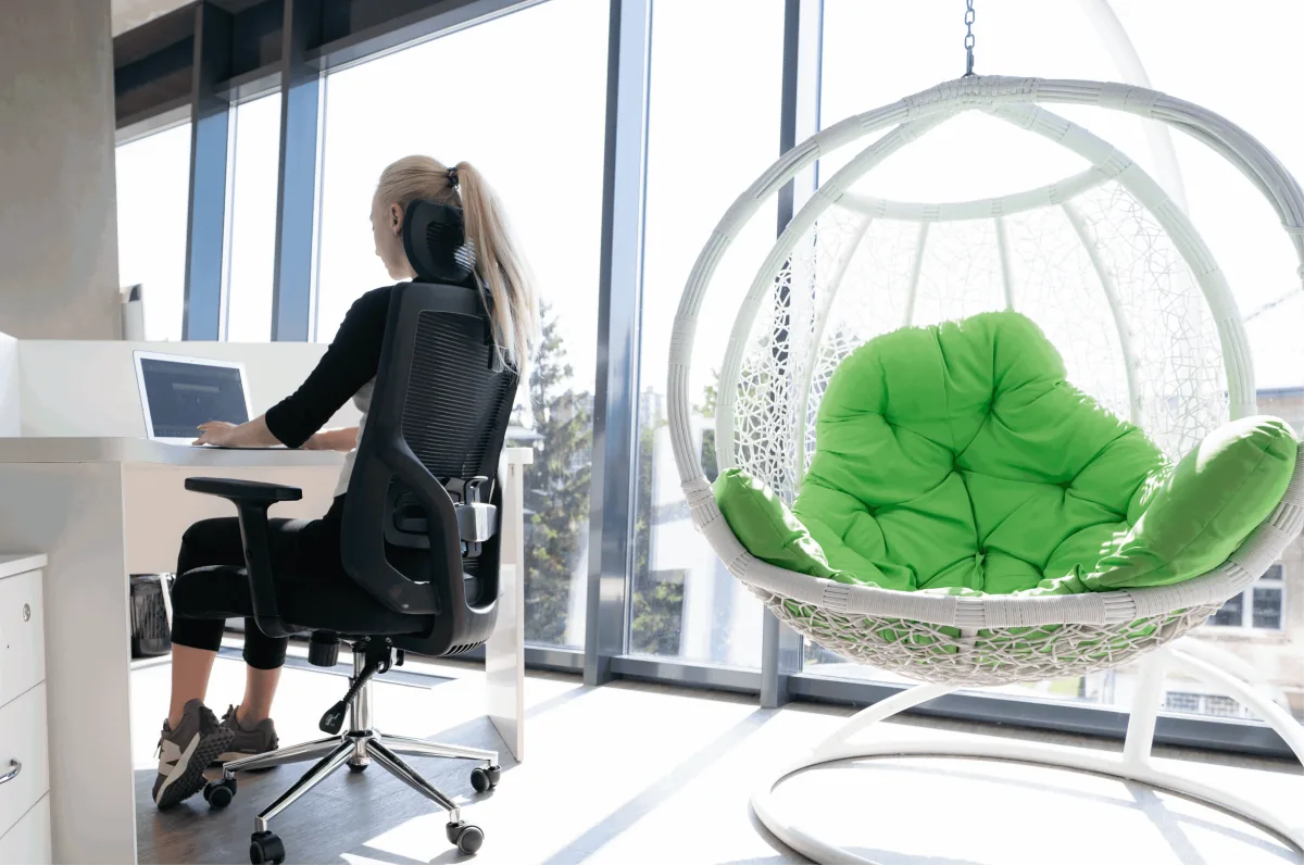 An employee works on a laptop at a window desk next to a hanging wicker egg chair with green cushions