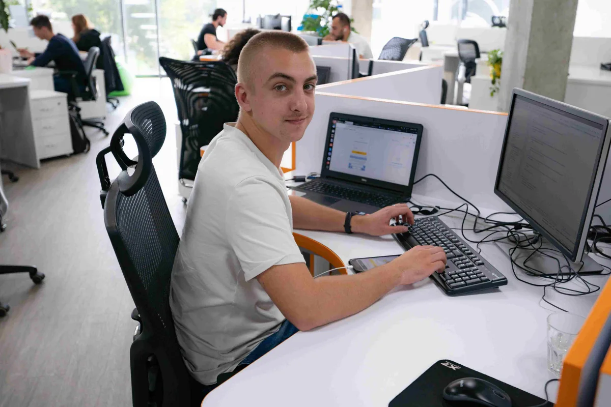 Young engineer smiles at the camera from his desk with a laptop and monitor in the open-plan office