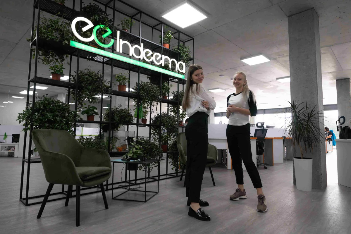 Two employees stand smiling by a plant shelf with an illuminated Indeema sign and a green armchair in the office lounge