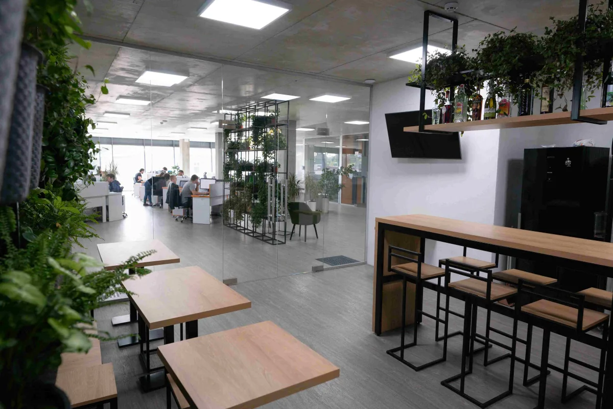 Office kitchen area with wooden tables and bar stools, hanging plants and a glass wall onto the open-plan workspace