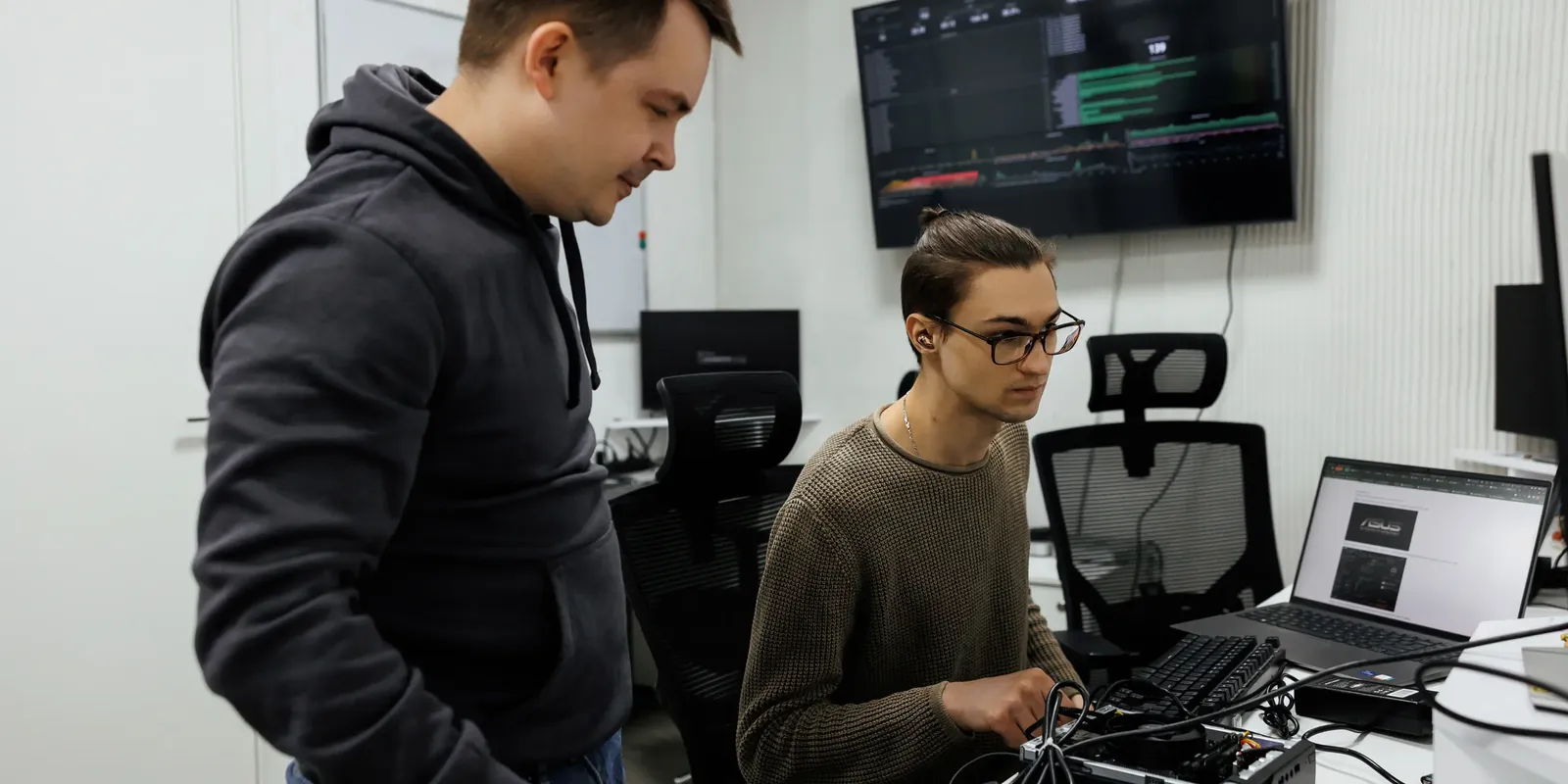 Two Indeema engineers at a test bench: one wires a connected hardware unit while a colleague watches, with live telemetry on a wall screen behind them