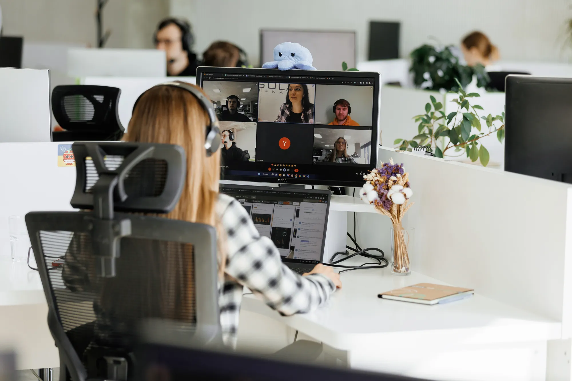 A person in headphones on a video call at an office desk, with colleagues working in the background