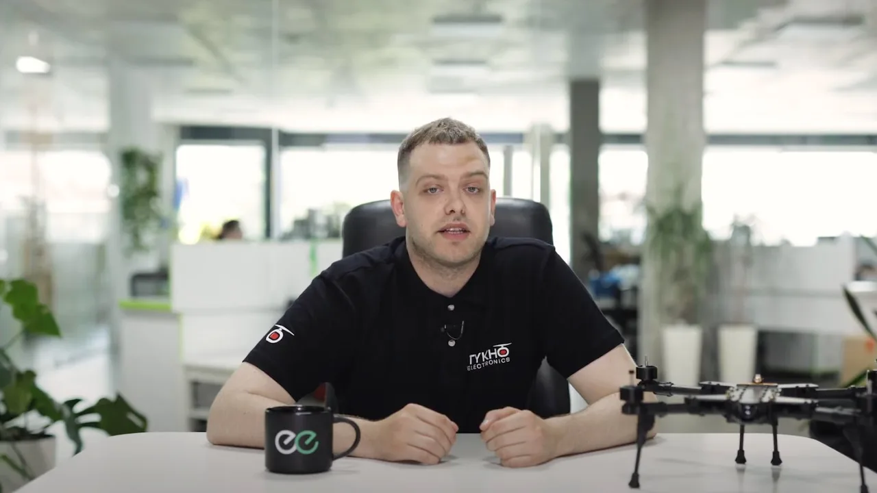 Video still: a man in a Tykho Electronics shirt speaking at a desk with an Indeema mug and a drone frame beside him